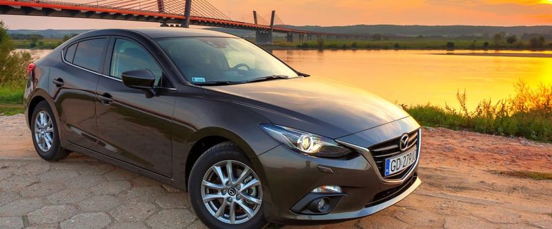 Image of a dark gray Mazda 3, parked outside with a sunset background and alongside the bridge and lake.