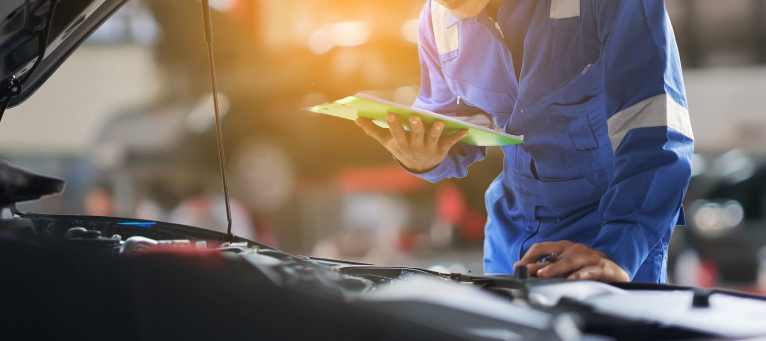 Image of a mechanic doing a inspection under the hood of a vehicle, with a clipboard.